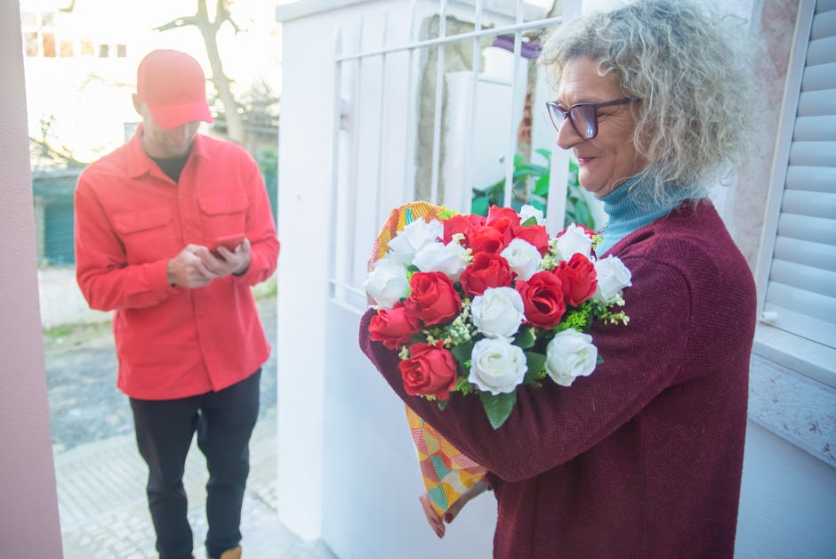 A woman with curly gray hair, wearing glasses, a red cardigan, and a blue turtleneck, holds a vibrant bouquet of fresh red and white roses wrapped in colorful paper, positioned in front of her chest,