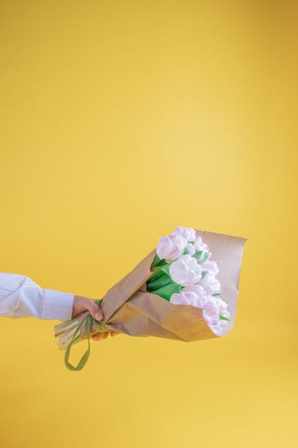 A person in a white shirt holds a bouquet of pale pink tulips wrapped in light brown kraft paper with a green ribbon, presenting a floral gift against a vibrant yellow background. The tulips have full