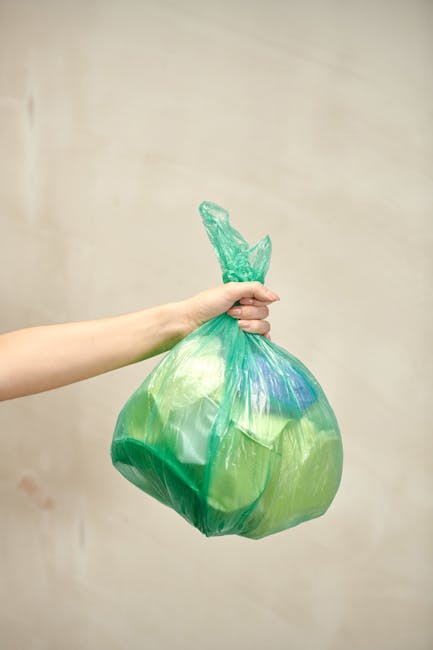 A person's arm holding a plastic bag filled with green waste, likely garden or floral debris, against a plain, light-colored background. The bag is tied at the top with a knot, and the waste inside ap