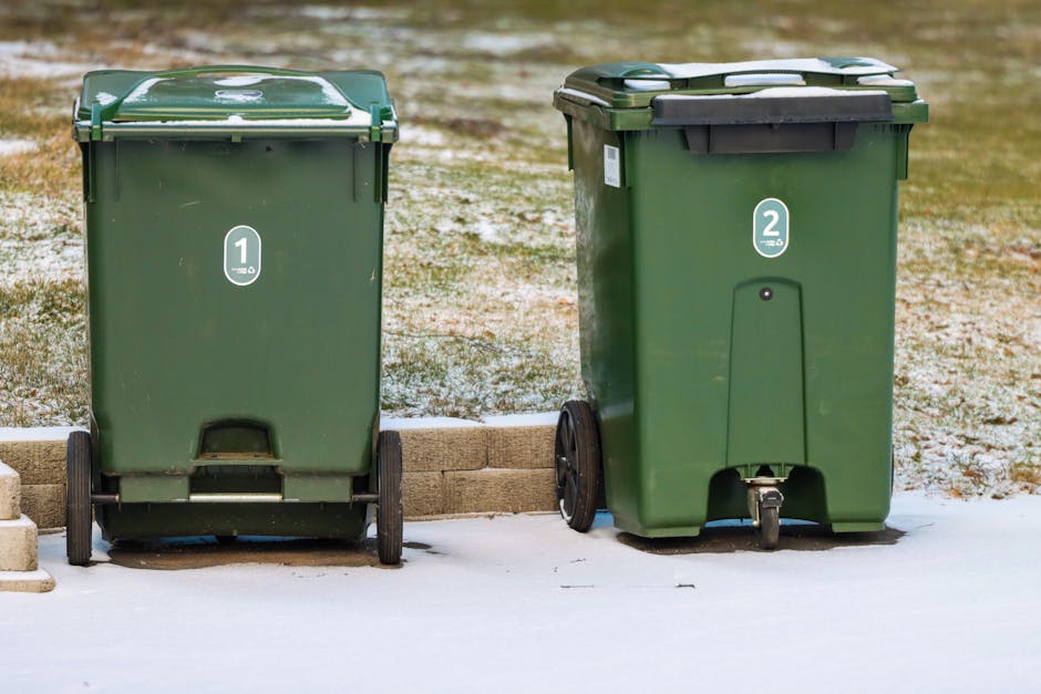 Two green wheeled trash bins labeled '1' and '2' are positioned outdoors on a snowy ground, with a low brick border separating them from a patch of grass and a paved area in the background. The first