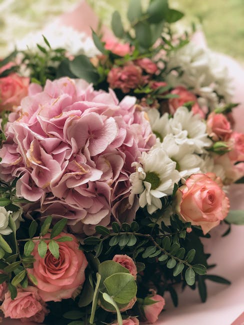 A floral arrangement featuring a large cluster of light pink hydrangea blooms with ruffled petals, surrounded by pale peach roses and small white daisies with yellow centers. The bouquet also includes