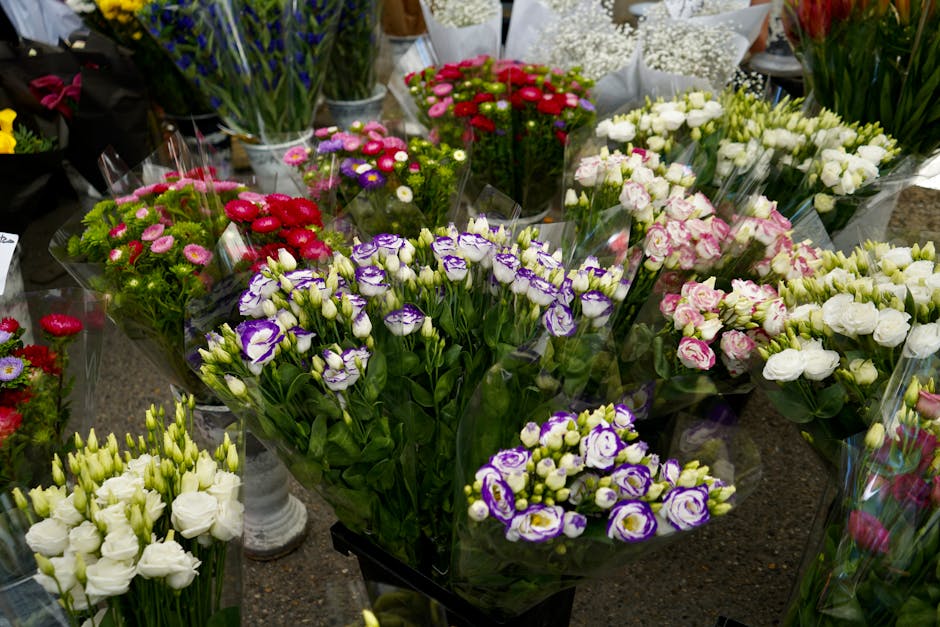 A vibrant display of fresh flower bouquets arranged on a dark surface inside a florist shop. The arrangement includes purple and white eustoma flowers, white lisianthus, pink and white roses, and red