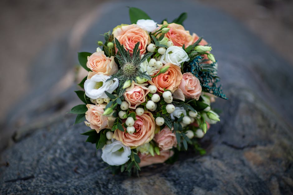 A wedding bouquet arranged with soft peach roses, complemented by white lisianthus, small white hypericum berries, and sprigs of blue thistles, all tied together with green foliage. The bouquet is res