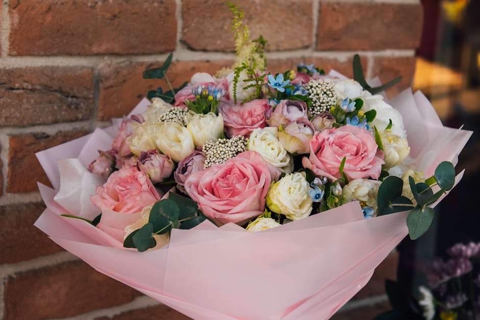 A floral bouquet arranged with pink roses, white lisianthus, small blue forget-me-nots, and sprigs of white baby's breath, accented with green eucalyptus leaves. The flowers are wrapped in pale pink t