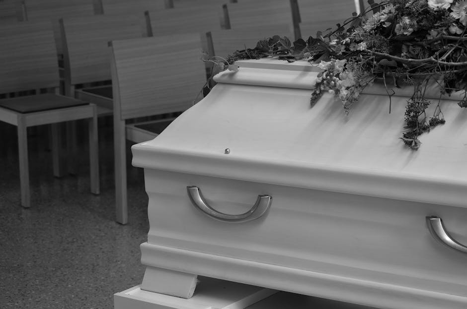 A white coffin with silver handles rests on a stand in an indoor setting, decorated with a lush arrangement of flowers and greenery draped across the top. The floral arrangement includes roses, lilies