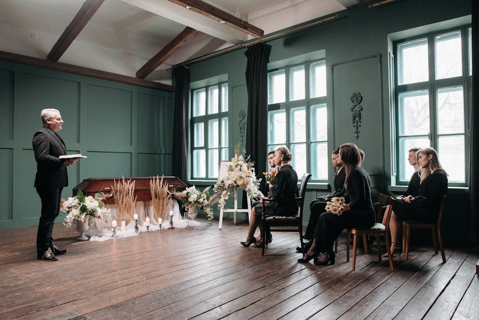 A floral arrangement consists of white roses, lilies, and carnations, accented with delicate greenery and dried wheat stalks, displayed on a white stand in front of a softly lit indoor setting. The fl